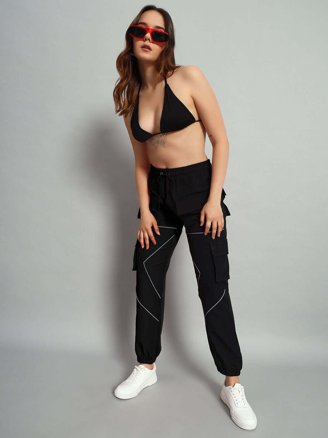 Model wearing WOMEN FRONTLINE REFLECTOR PARACHUTE CARGO PANTS BLACK with red sunglasses and white sneakers against a gray background.
