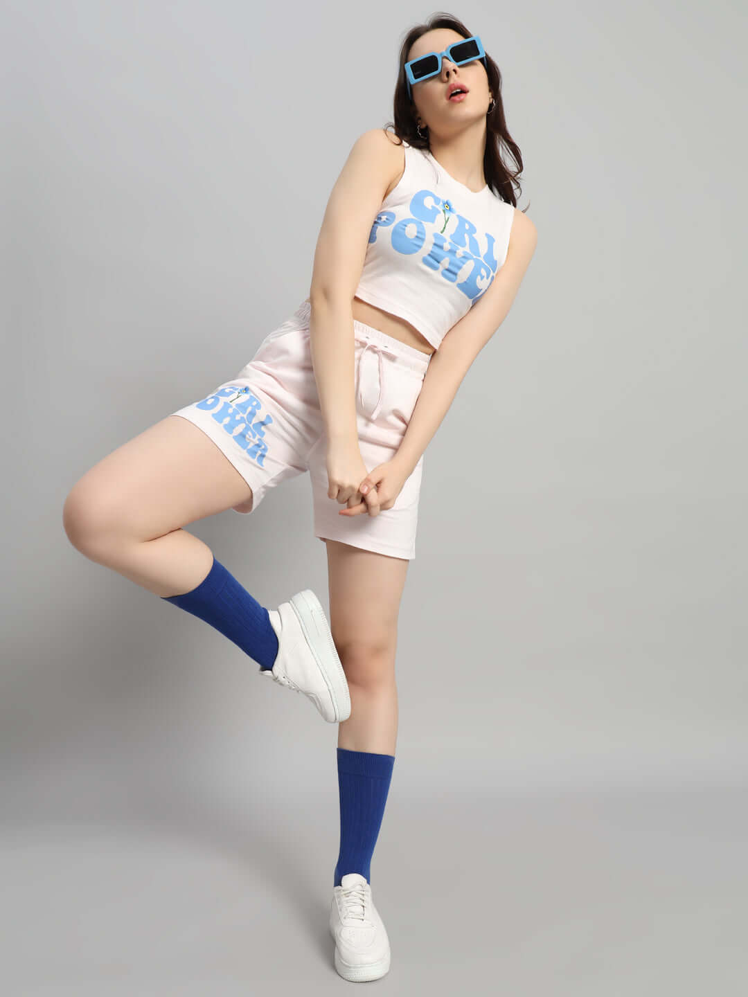 Woman in baby pink GIRL POWER co-ord set with blue print, posing confidently in sunglasses and sneakers against a gray background.