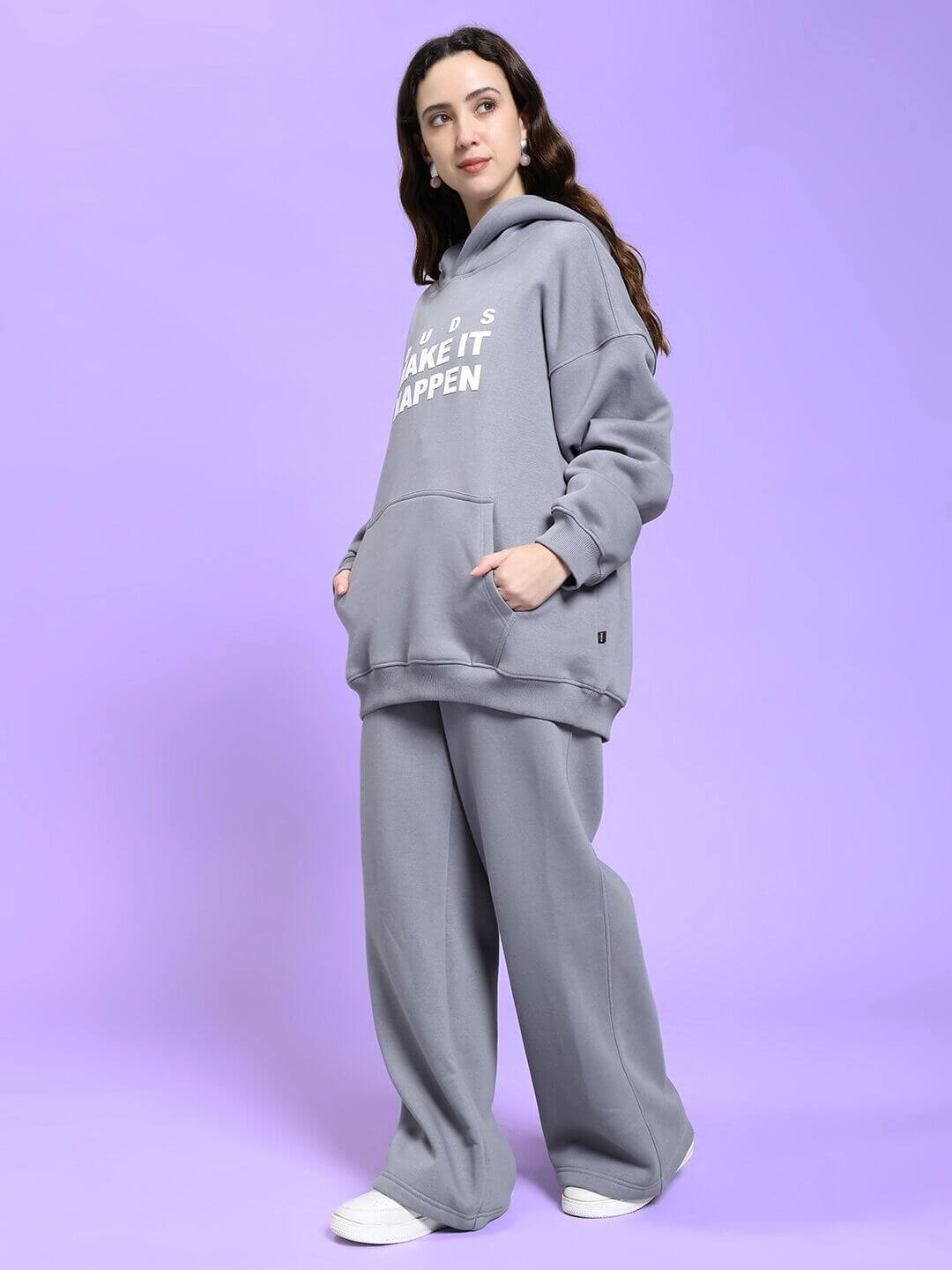 Woman wearing WOMEN MAKE IT HAPPEN FLEECE CO-ORD SET GREY, standing on a purple background, showcasing cozy streetwear style.