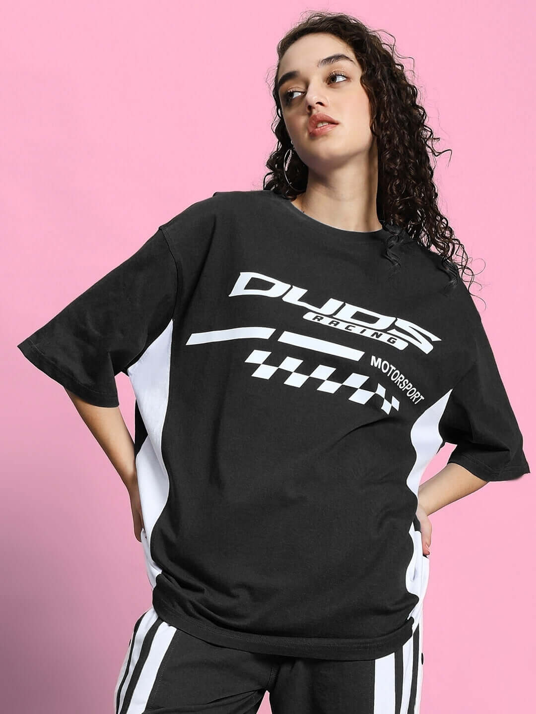 Woman wearing WOMEN'S INDY OVER-SIZED CLASSIC T-SHIRT BLACK with graphic design, featuring premium heavy gauge fabric on a pink background.