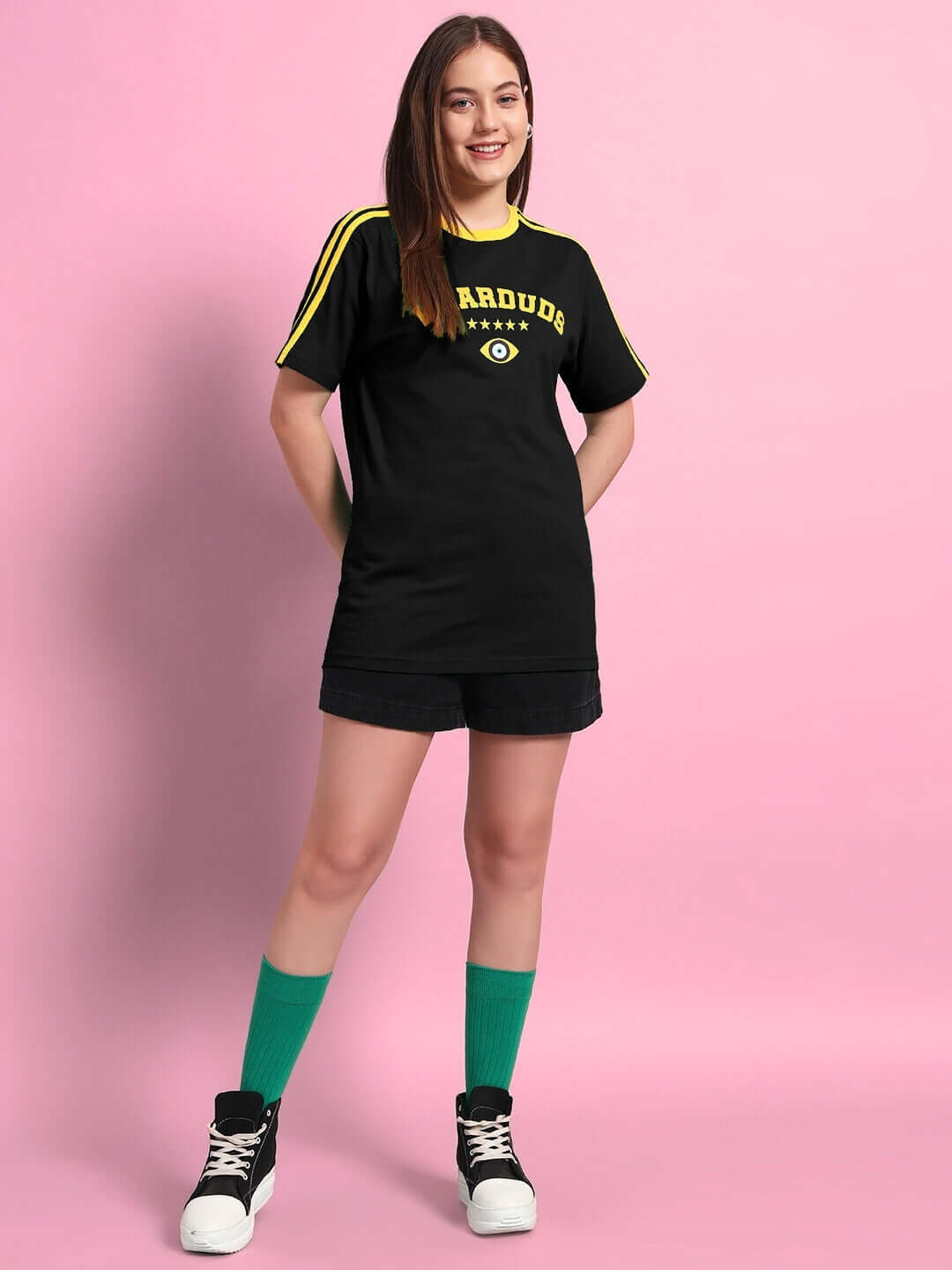 Model wearing women's black Irish T-shirt, premium cotton, with yellow accents, styled with green socks and sneakers on a pink background.