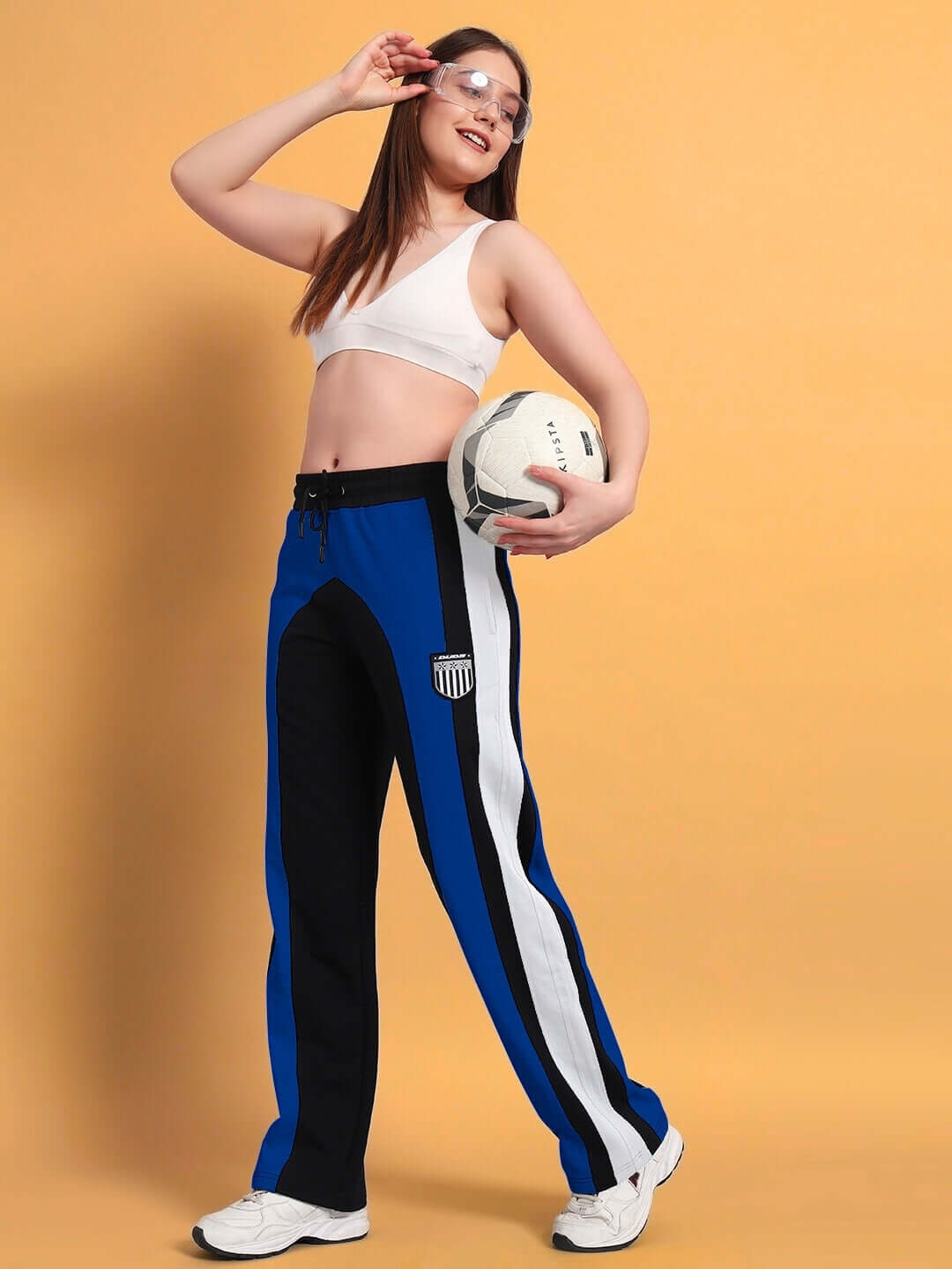 Woman wearing black and blue color block joggers holding a soccer ball, against an orange background.
