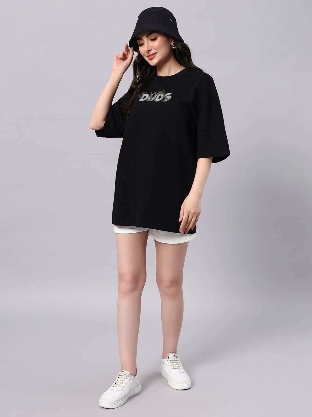 Model wearing a women's oversized black t-shirt, paired with a bucket hat and white sneakers, showcasing casual fashion style.