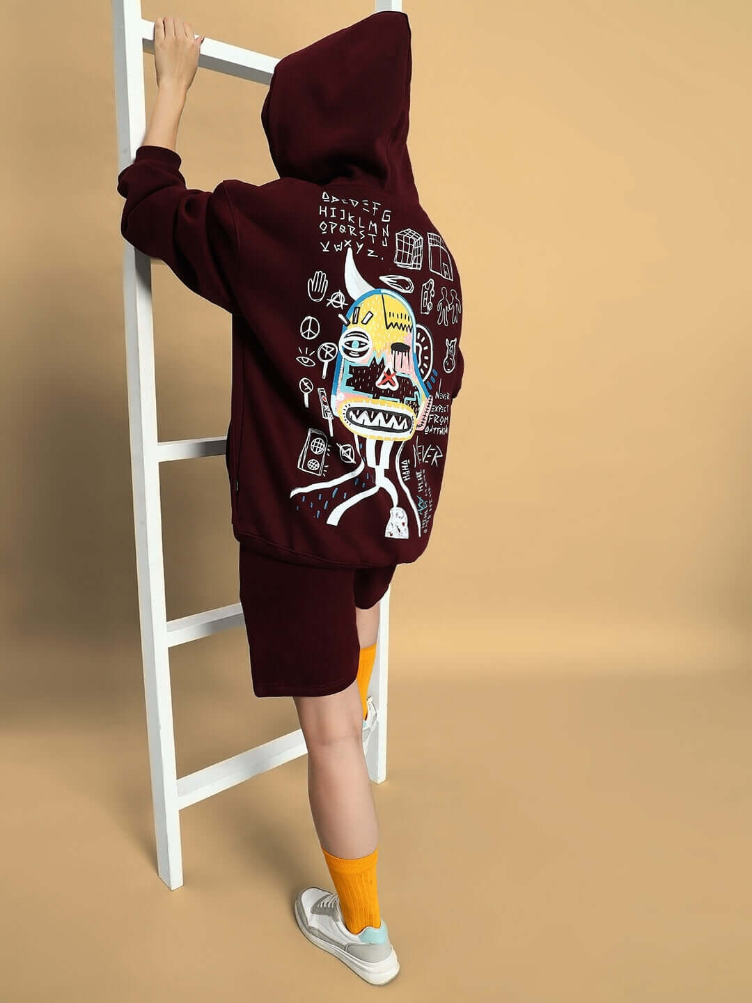 Woman wearing a wine-colored jumbled fleece co-ord with graphic design on the back, standing by a white ladder.