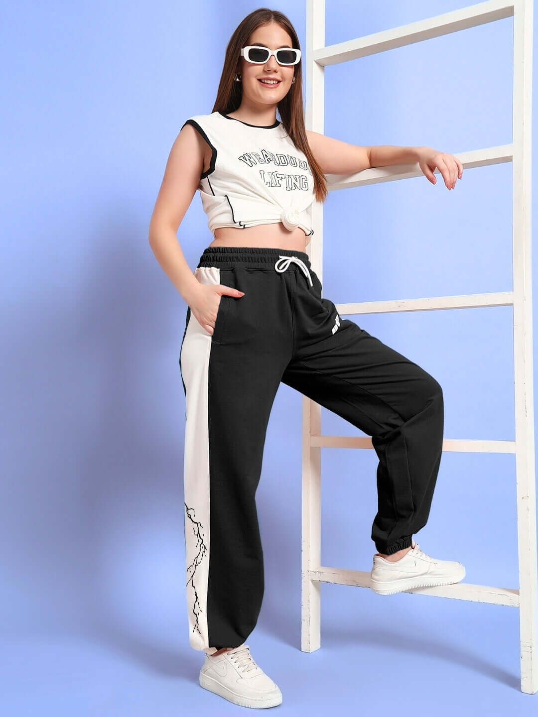 Woman wearing black-white relaxed fit joggers with lightning bolt design, posing stylishly with sunglasses on a blue background.