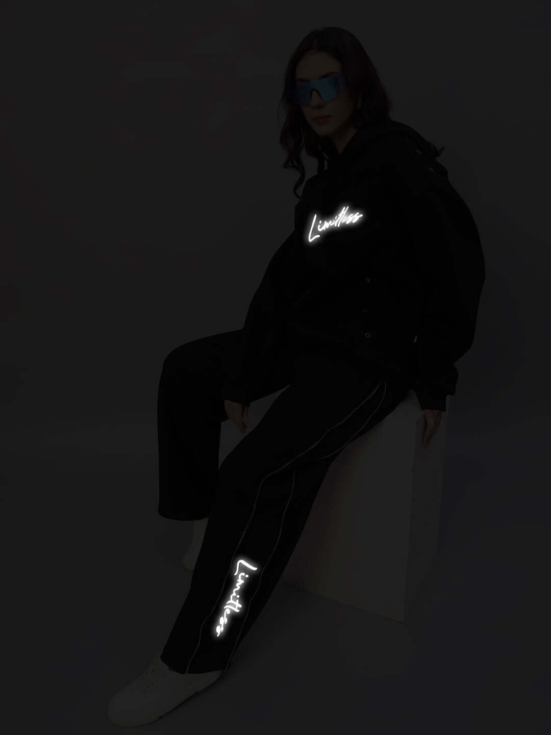 Model wearing Women's Limitless Fleece Reflector Co-Ord in Black with reflective details, sitting on a white block against a dark background.