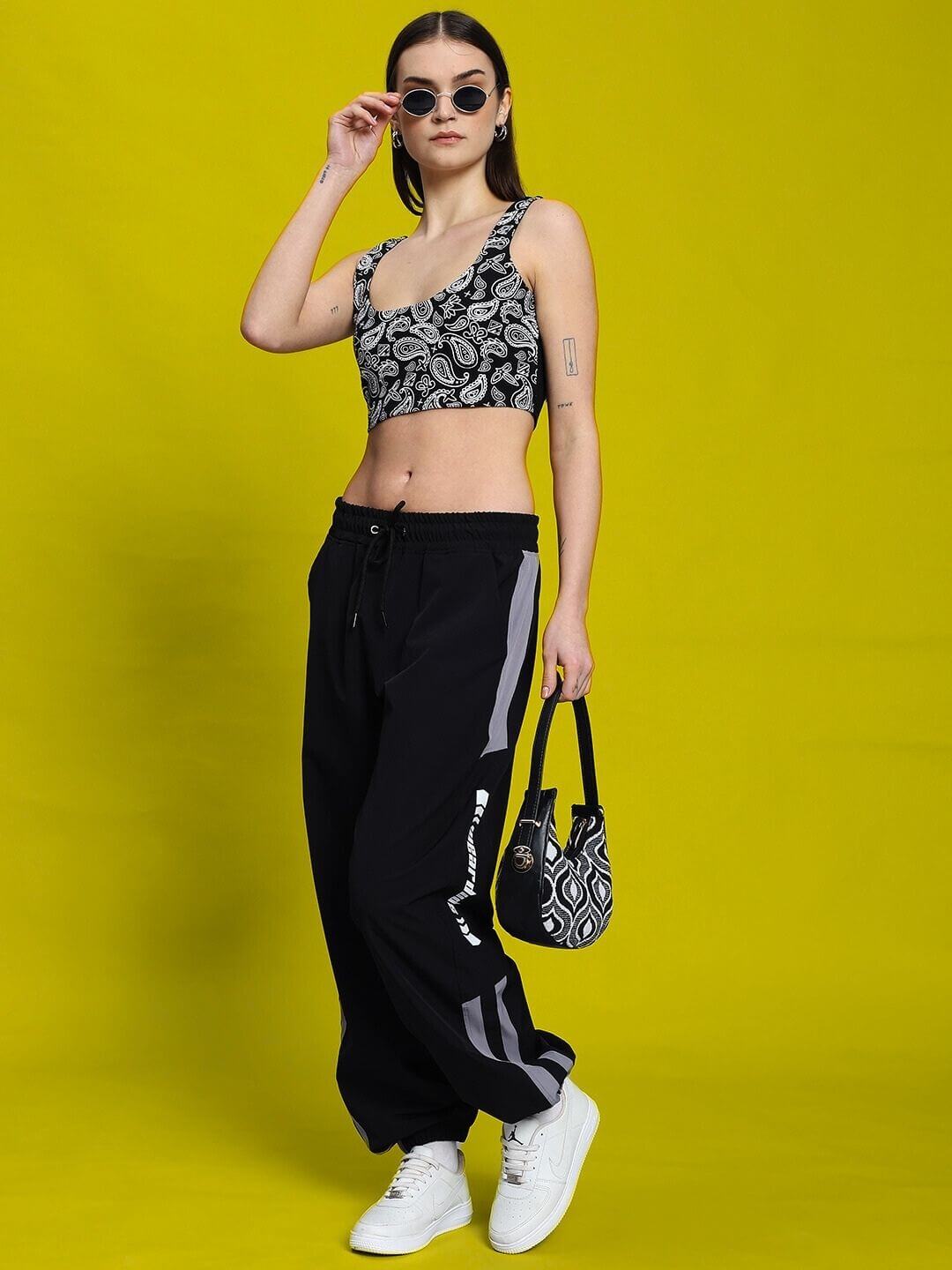 Model posing in WOMEN'S LINNA CO-ORD SET BLACK by WEAR DUDS in a stylish fashion look with sunglasses and purse on a yellow background.