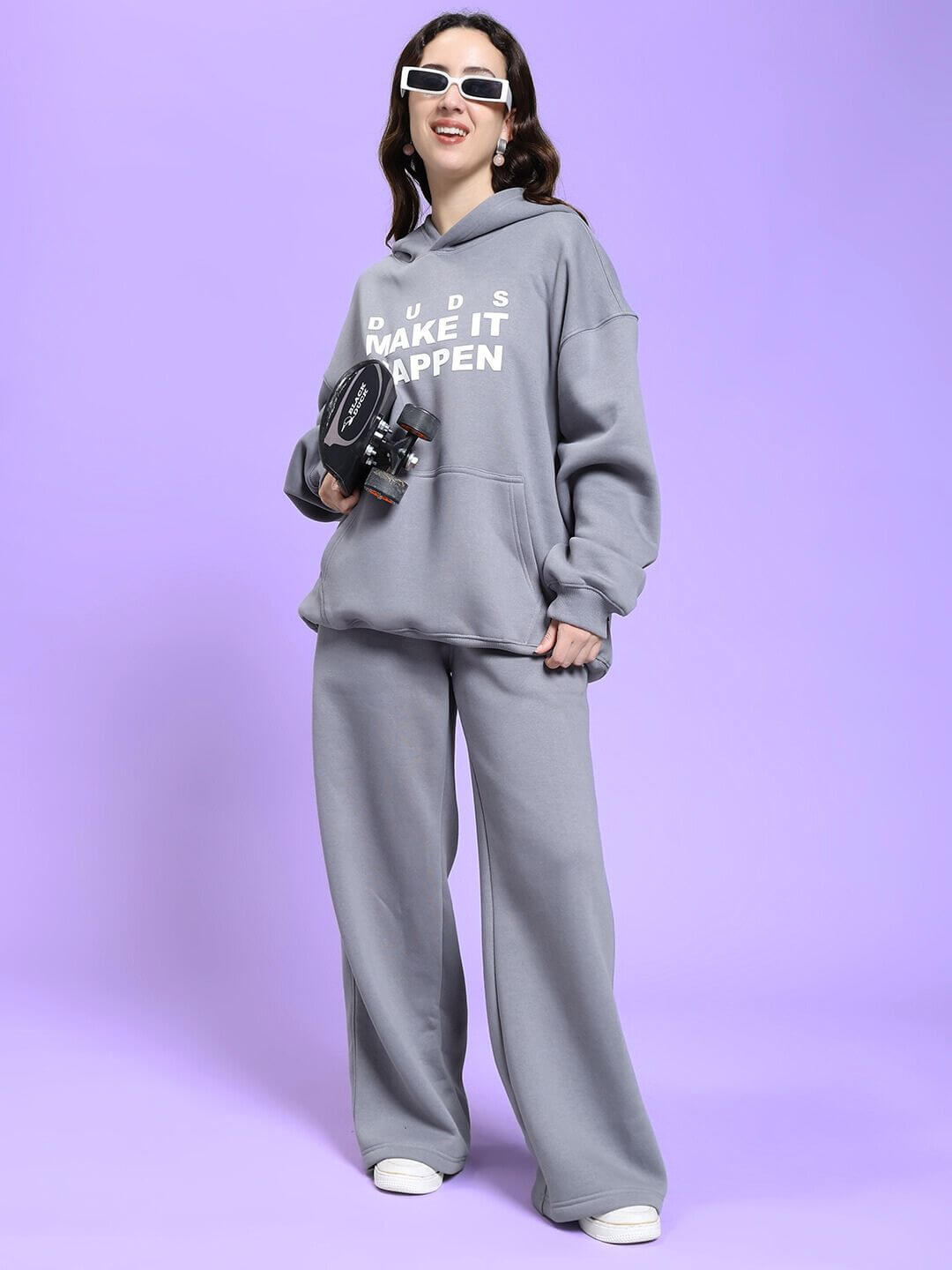 Woman wearing WOMEN MAKE IT HAPPEN FLEECE CO-ORD SET GREY, holding a camera, radiating casual streetwear style against a purple background.