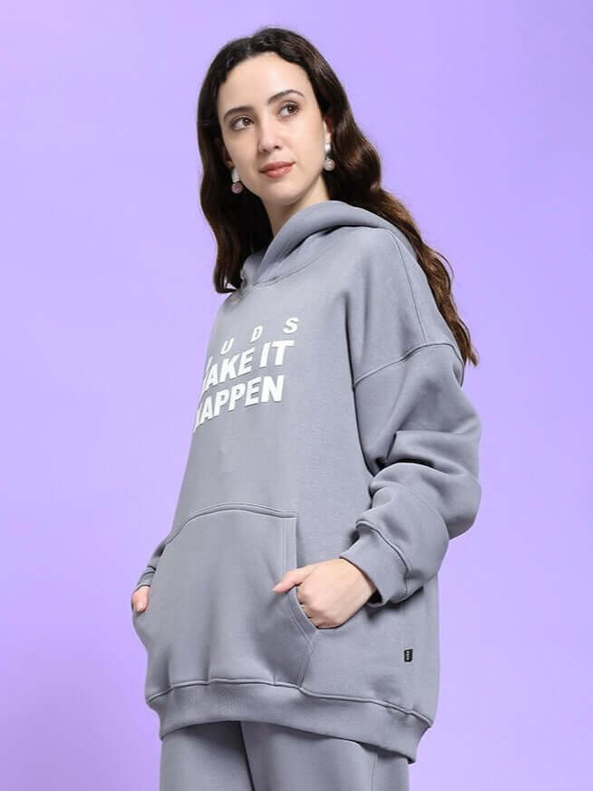 Model wearing WOMEN'S MAKE IT HAPPEN FLEECE HOODIE GREY with hands in pockets, against purple background.