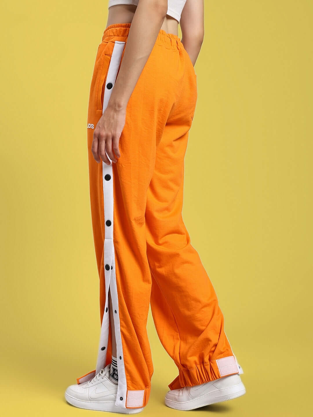 Woman wearing orange relaxed fit joggers with side button detail, standing against a yellow background.