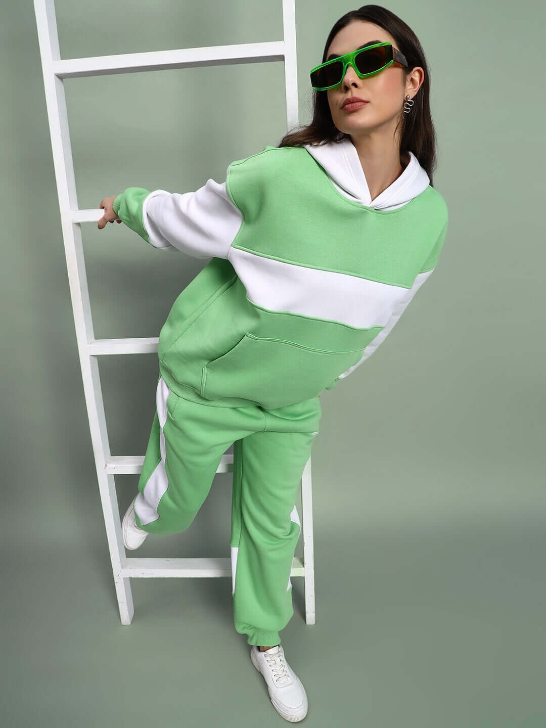 Model wearing WOMEN MINTED BLISS FLEECE CARGO PANT GREEN WHITE with matching hoodie and sunglasses, leaning on a white ladder.