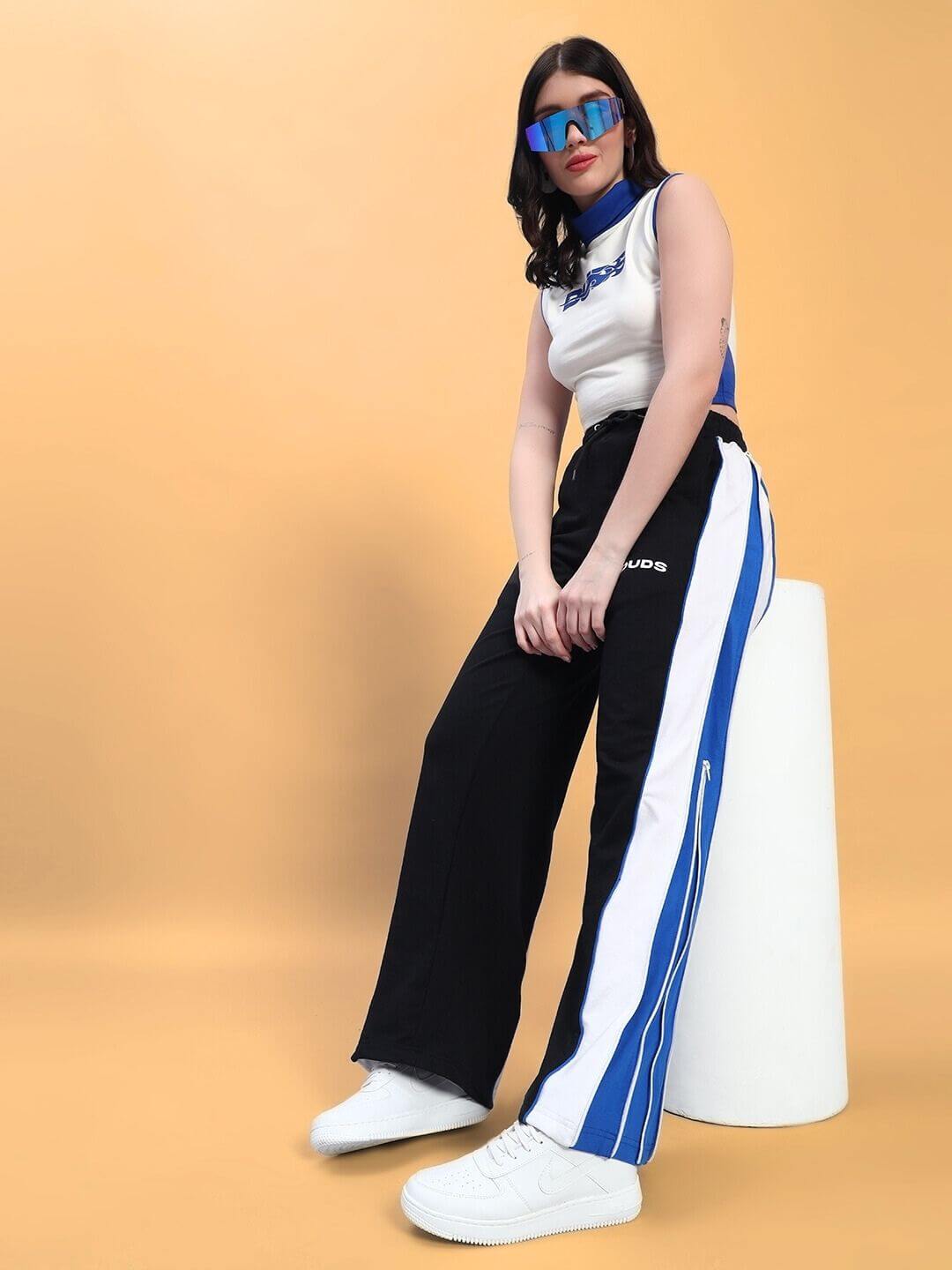 Woman wearing stylish sporty outfit with a white crop-top and blue-striped black pants, sitting on a pedestal against a peach background.