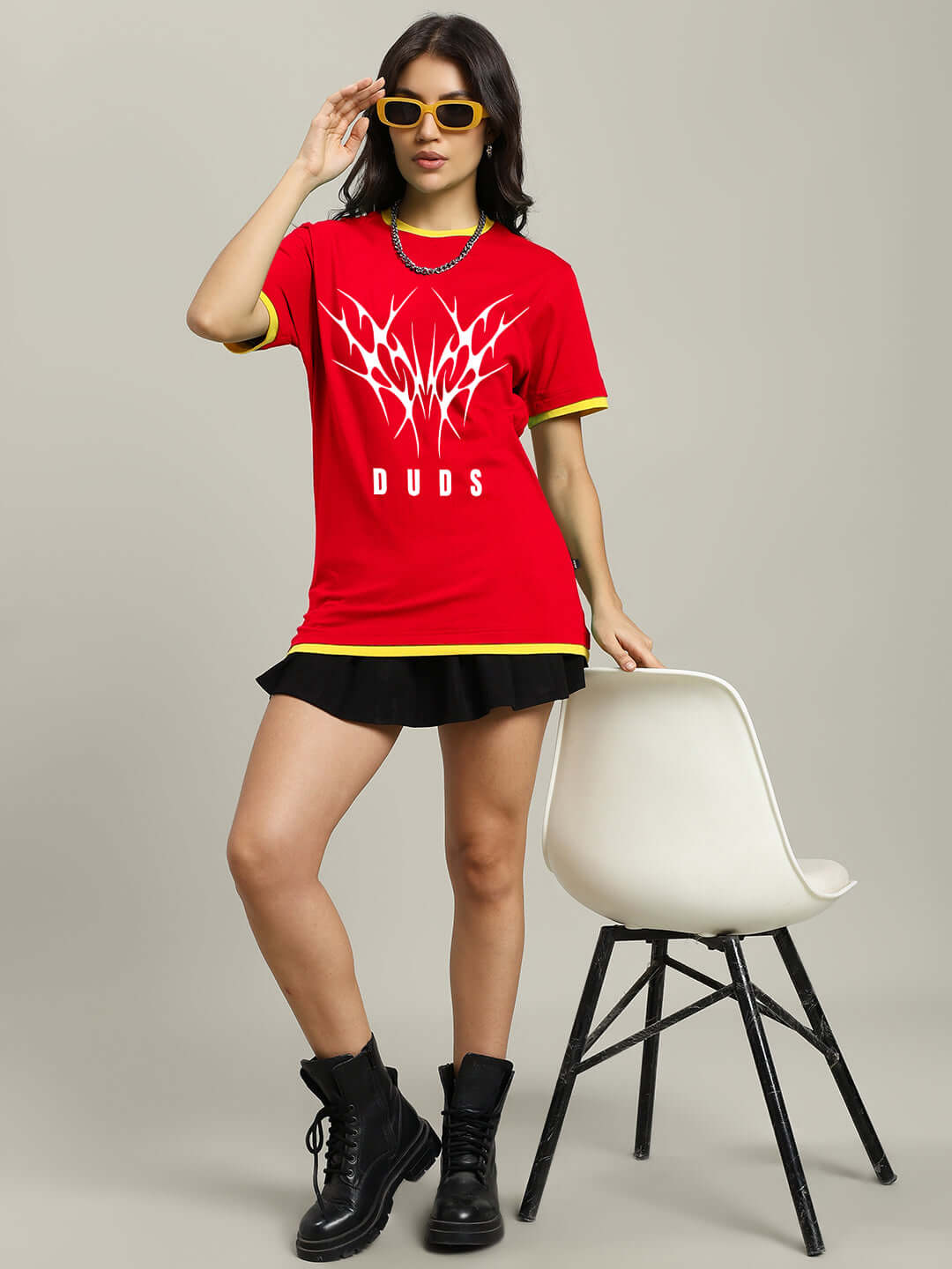 Woman wearing a red Noriba regular fit T-shirt with unique graphic, paired with black skirt and boots, stands beside a white chair.