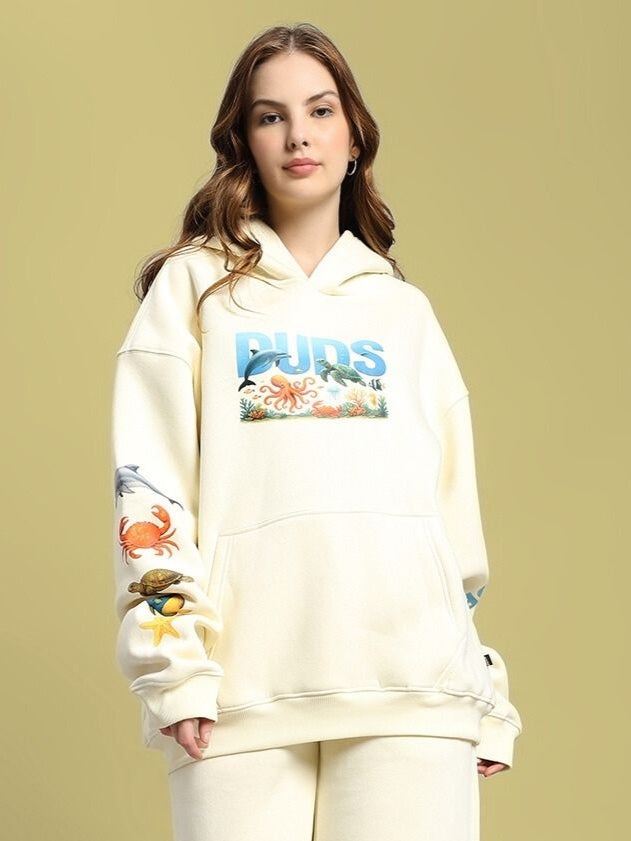 Model wearing WOMEN'S OCEAN BUDDIES OVERSIZED FLEECE HOODIE OFF-WHITE with ocean-themed graphics on sleeves and chest.