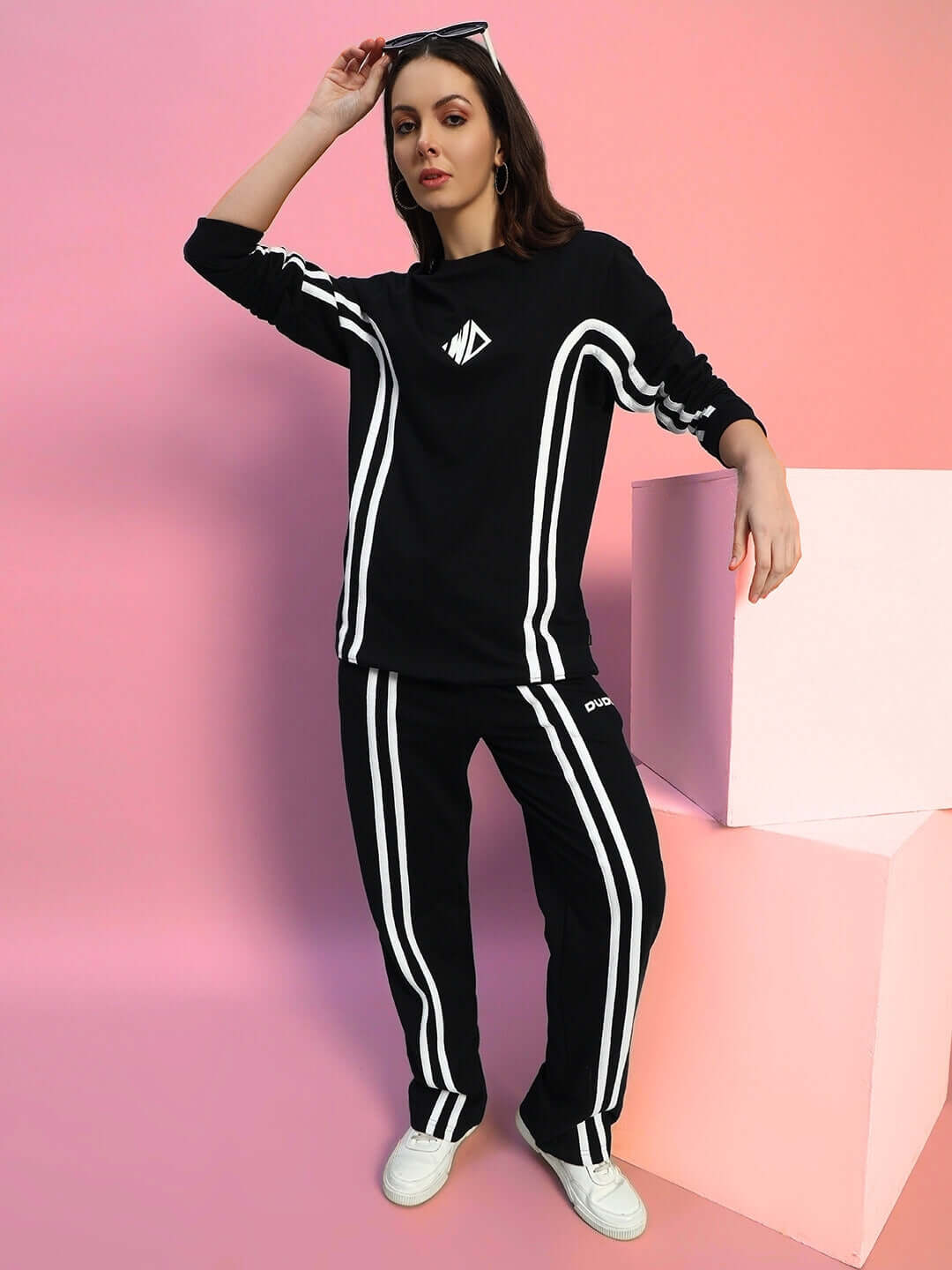 Woman wearing black women's jogger set with white stripes, styled with white sneakers, posed against a pink background.