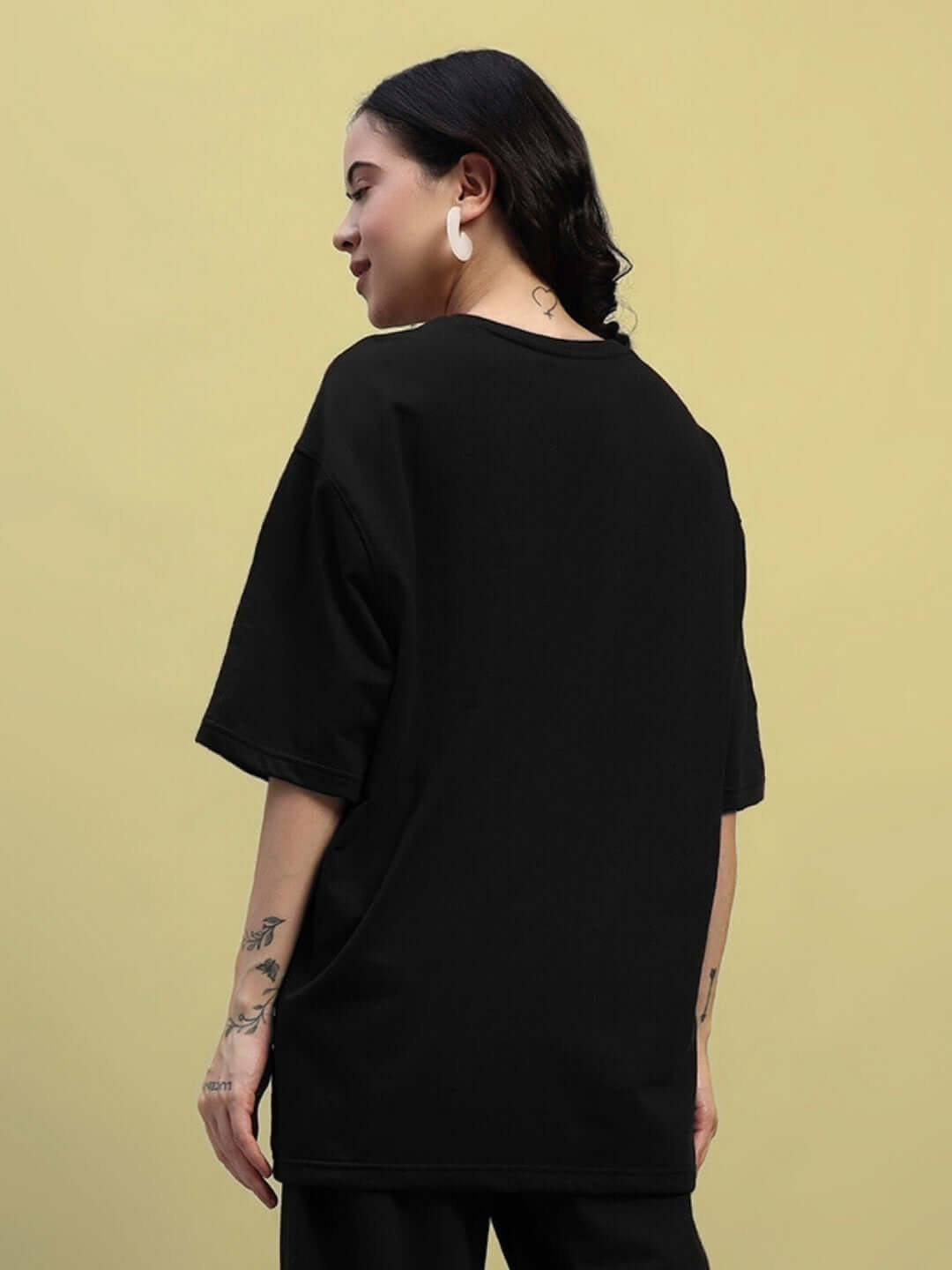 Back view of a woman wearing a black oversized t-shirt made of premium cotton fabric with tattoos visible on her arms.