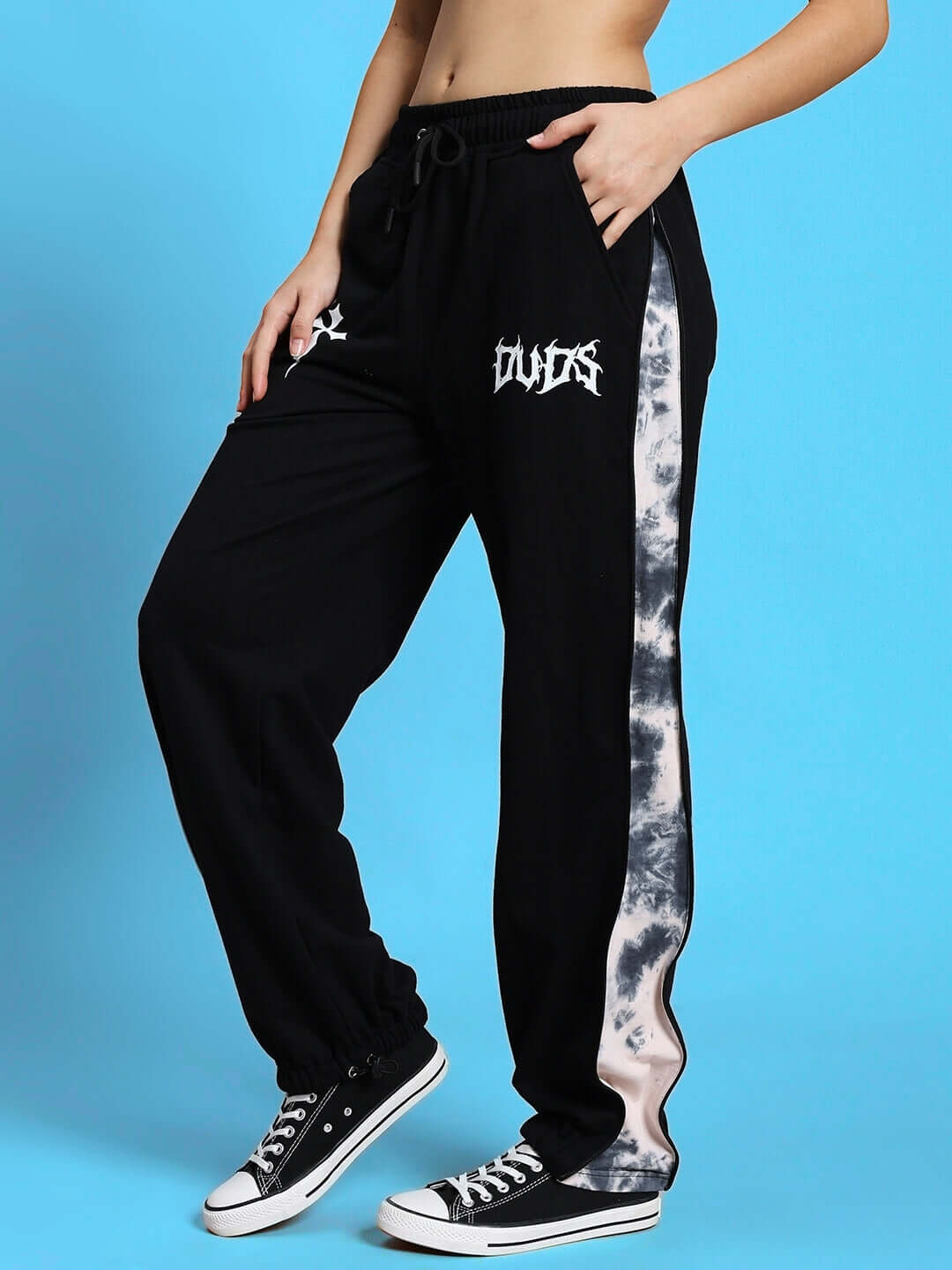Model wearing black joggers with zipper panels, featuring tie-dye side strips and logo, paired with sneakers against a blue background.