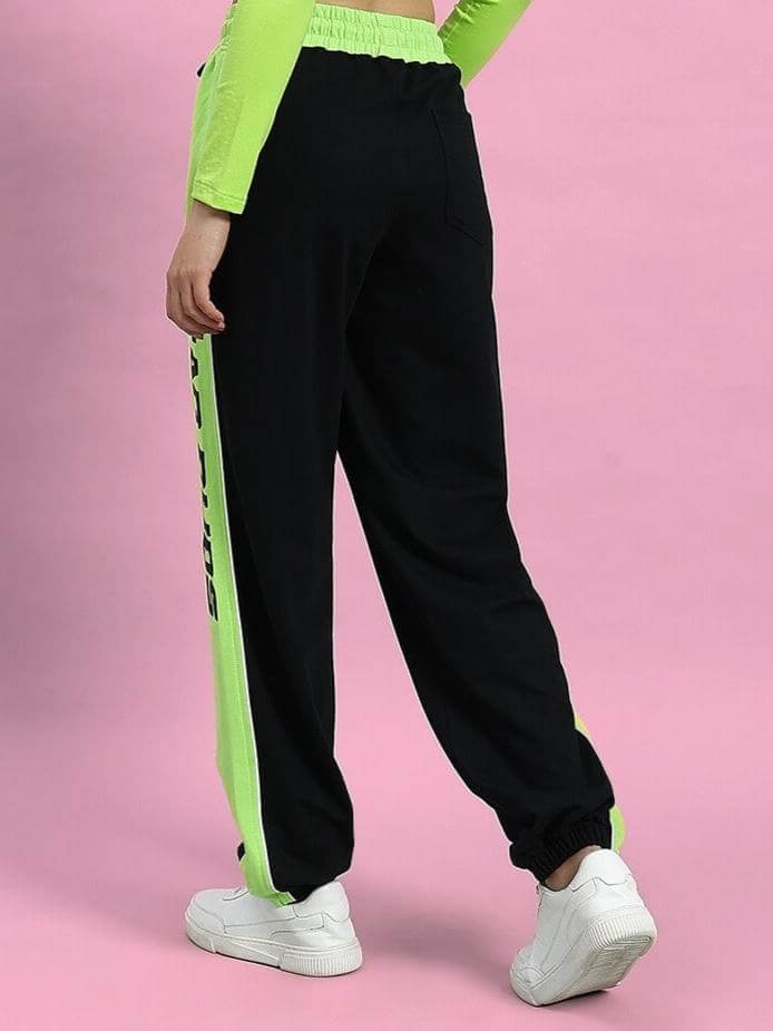Back view of Women's Peppy Joggers in Black and Neon Green with side stripe detail, worn with a neon green top against pink background.
