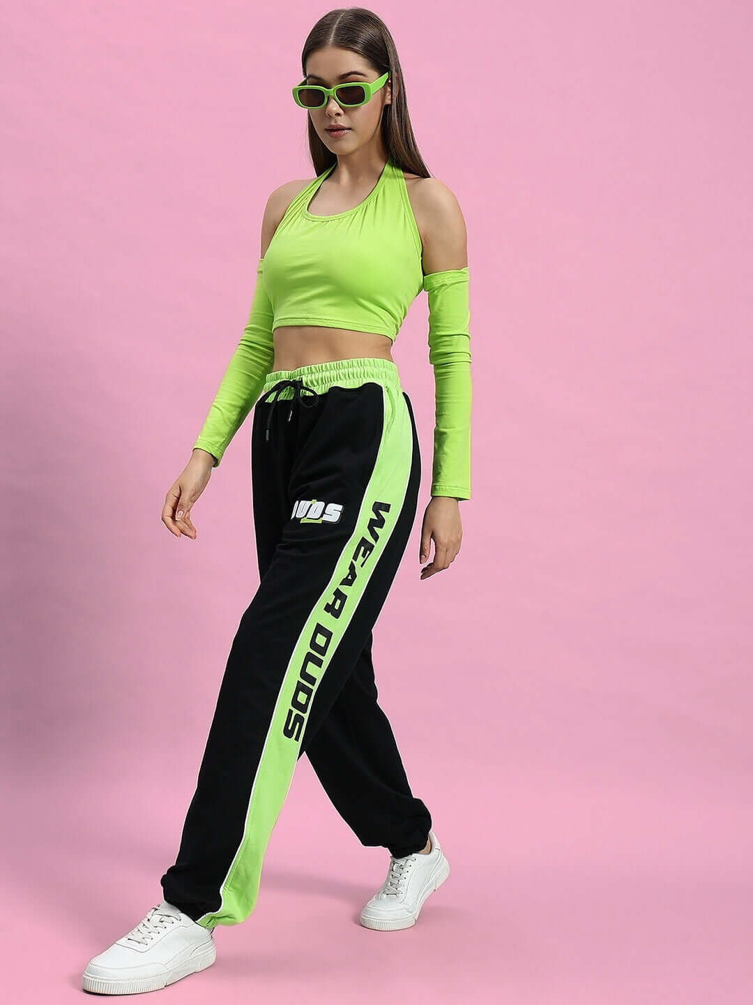 Woman wearing black-neon green joggers with "Wear Duds" branding, styled with matching neon top and sunglasses, against pink background.