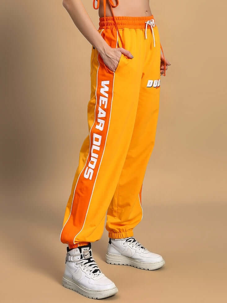 Women's Peppy Joggers in yellow-orange by DUDS, featuring side stripes and elastic waist, showcased by a model standing.