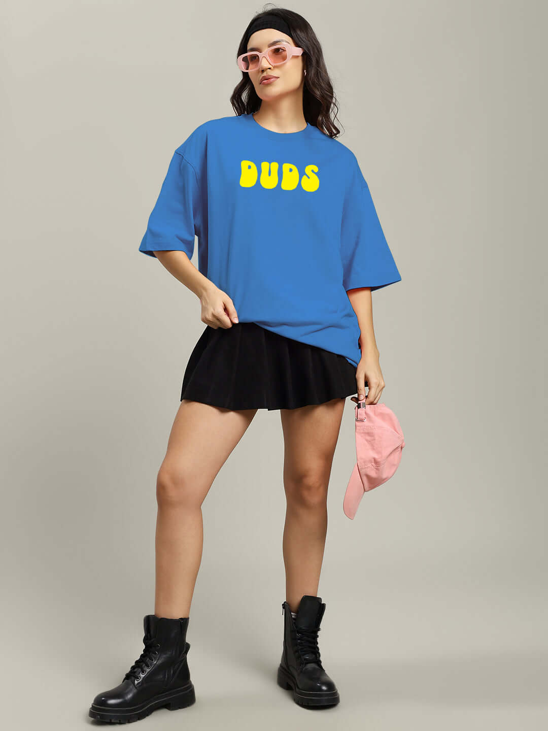 Model wearing WOMEN'S PERSONAL TRUTH oversized blue T-shirt with 'Duds' text, styled with black boots and skirt, holding pink cap.