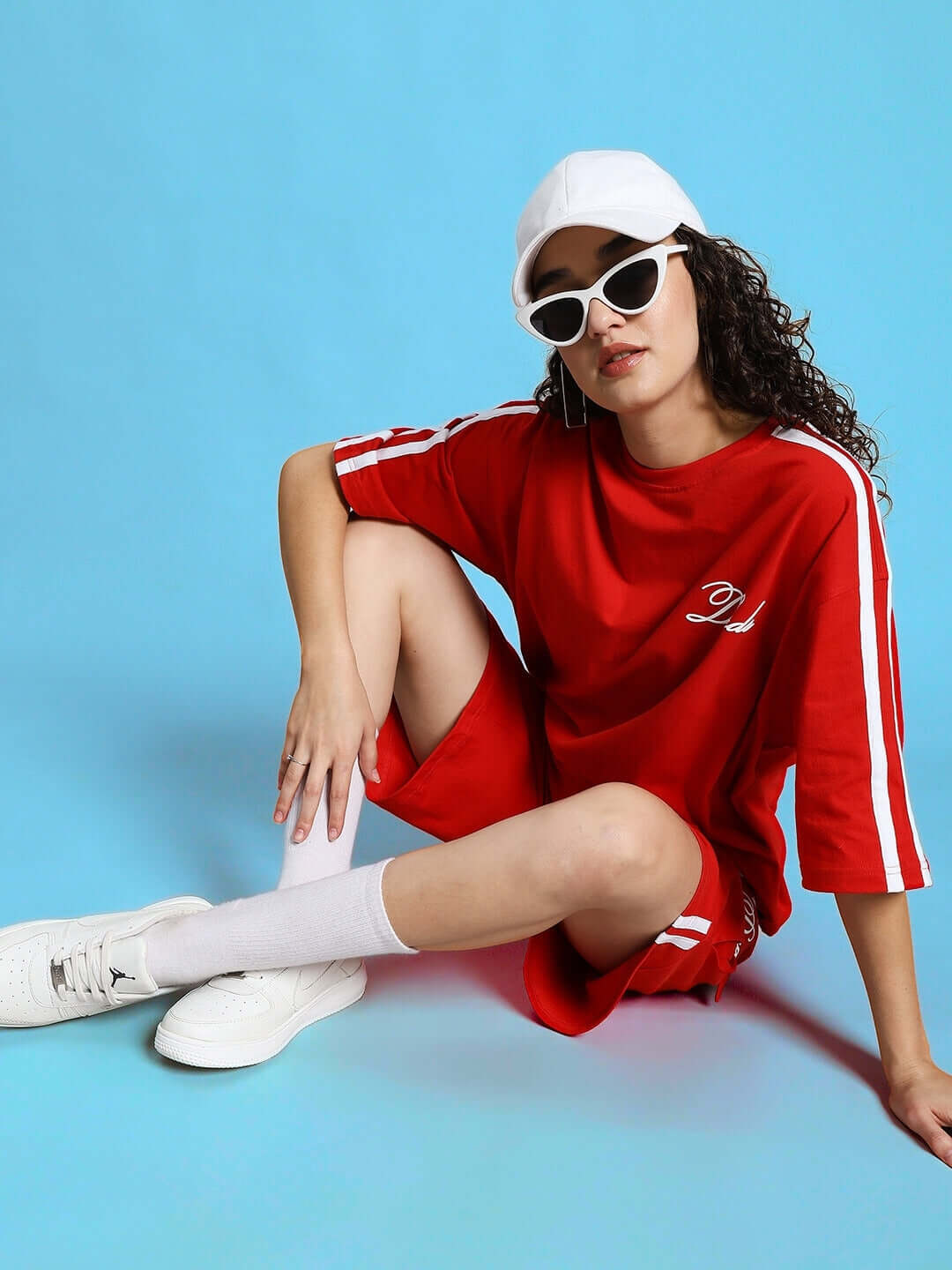 Woman wearing red Women's Scotia Regular Fit Shorts with matching top, white sneakers, and hat, sitting against a blue background.