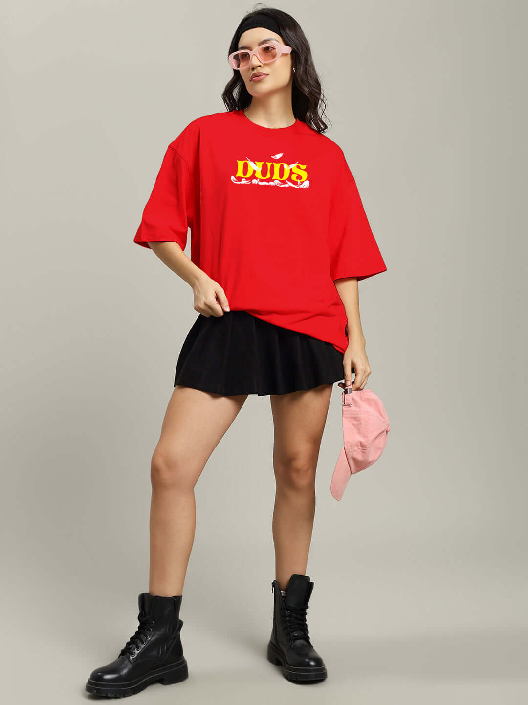Woman wearing red oversized T-shirt with "Stay Happy" print, black skirt, pink cap in hand, and black boots, styled with sunglasses.