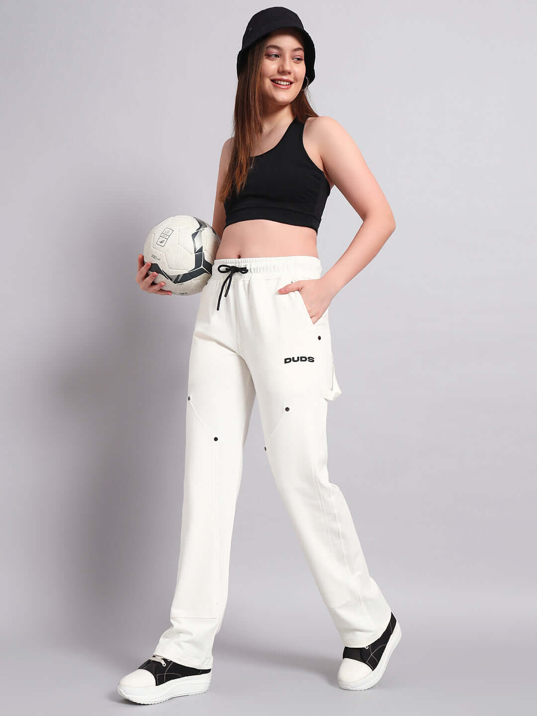 Model wearing white women's Regulus relaxed-fit joggers by DUDS, styled with a black tank top and holding a soccer ball.