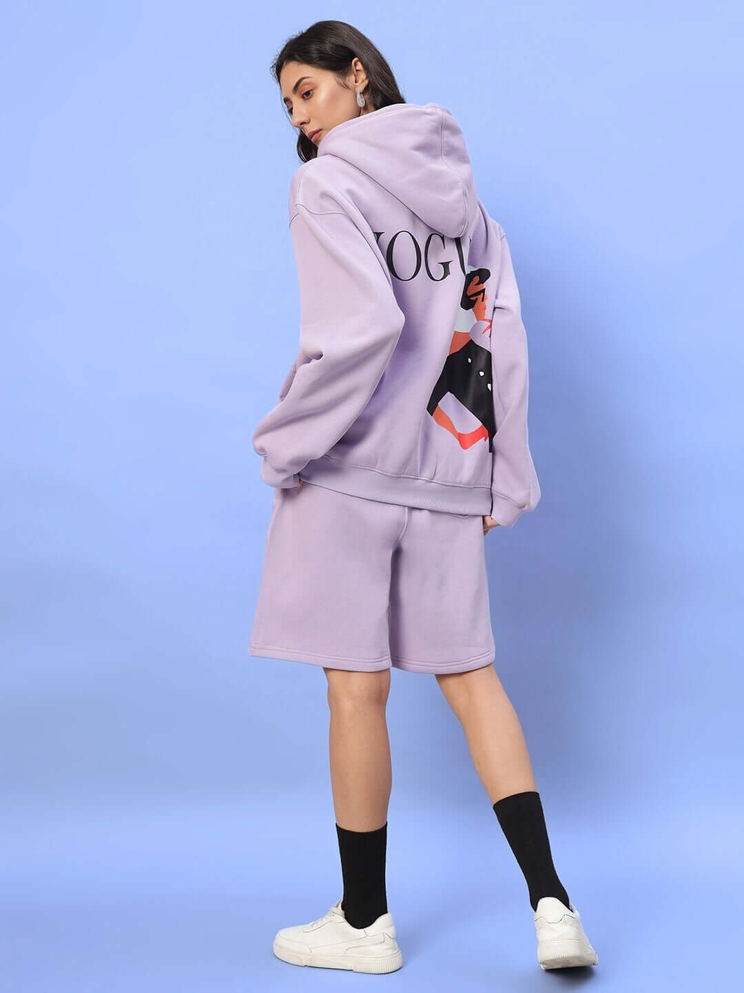 Woman wearing WOMEN RITZY FLEECE CO-ORD LILAC set with graphic hoodie, shown from the back, against a blue background.