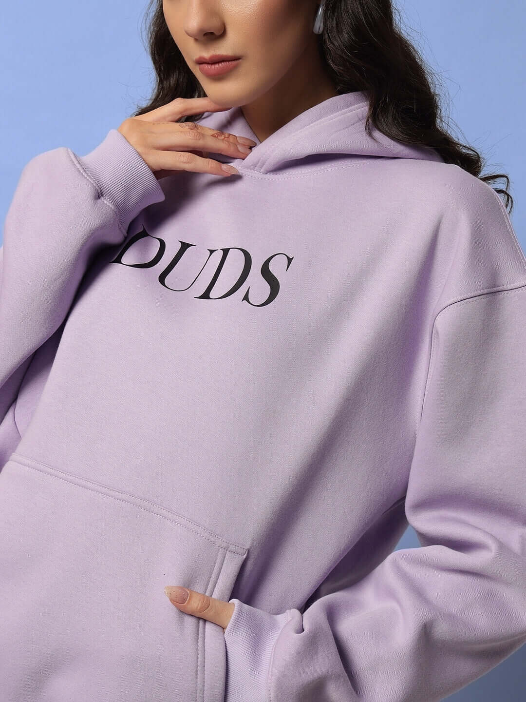 Woman wearing WOMEN RITZY FLEECE HOODIE LILAC in soft lilac color with "DUDS" printed on front, showcasing style and comfort.