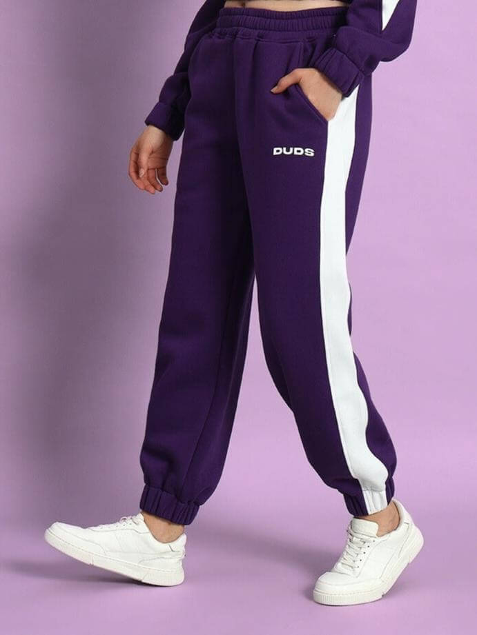 Woman wearing WOMEN RIZZLER FLEECE COLORBLOCK JOGGER PURPLE with white sneakers standing against a lavender background.