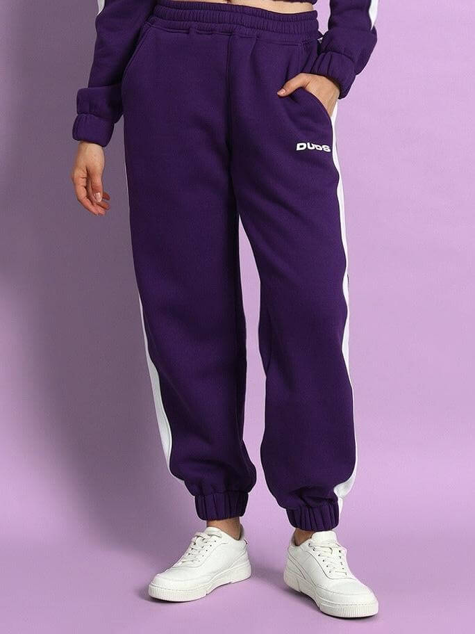 Model wearing WOMEN RIZZLER FLEECE COLORBLOCK JOGGER PURPLE, showcasing stylish purple joggers with white side stripes and sneakers.