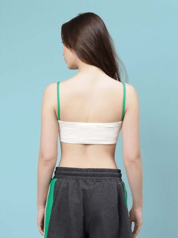 Back view of a woman wearing an off-white crop-top with green straps, styled with gray sporty pants, against a light blue background.