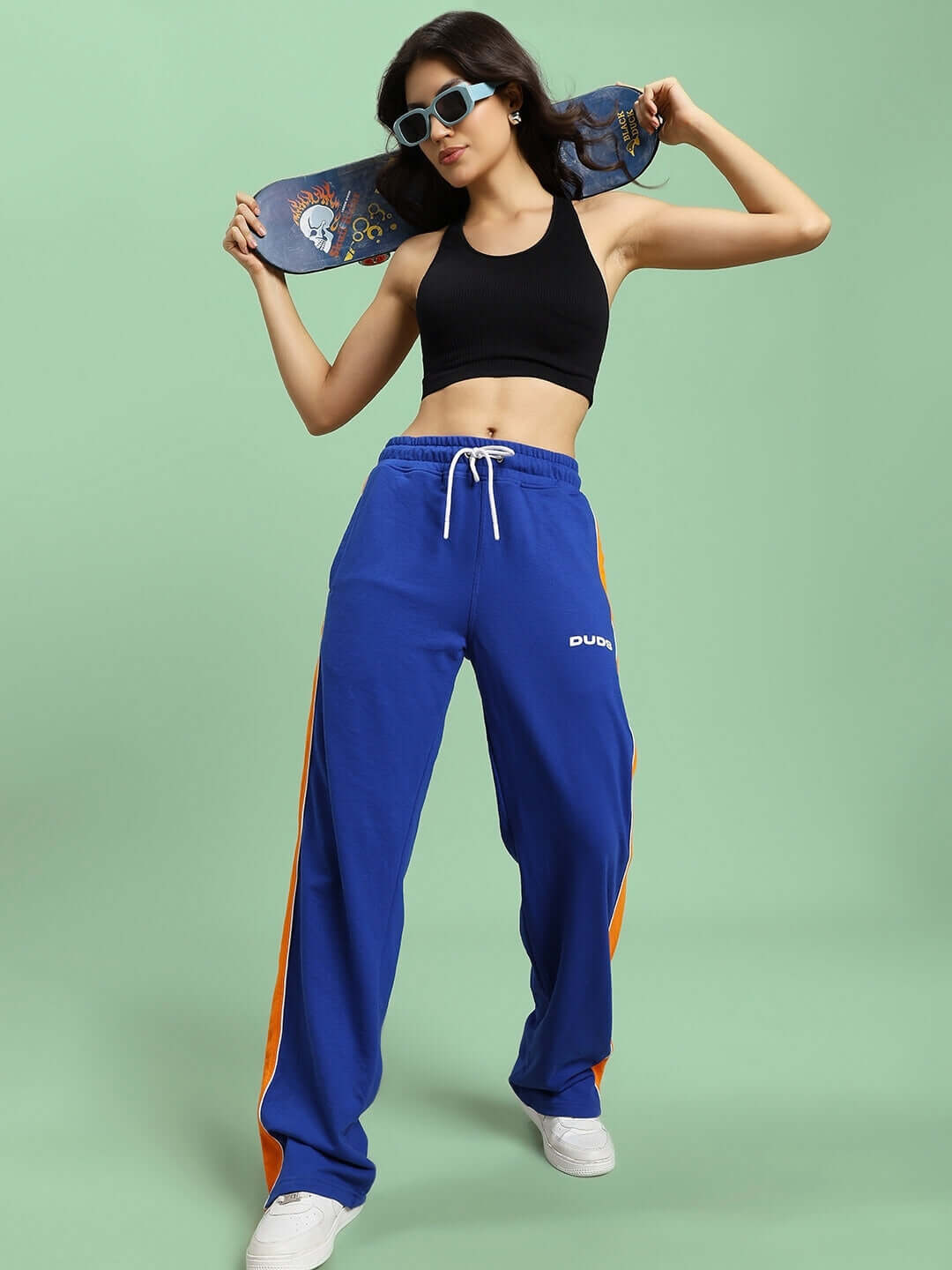 Woman wearing WOMEN'S SANFORD STRIPE JOGGERS BLUE with a black top, posing confidently with a skateboard on a green background.