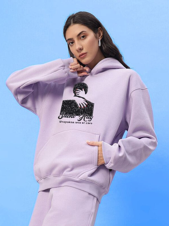 Woman wearing WOMENS SILENT HUG PRINTED FLEECE HOODIE LILAC with graphic design on chest, against blue background.