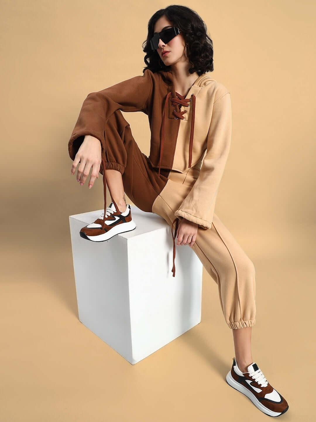 Woman wearing a stylish beige-brown co-ord set, sitting on a white cube, showcasing chic fashion and comfort with sneakers and sunglasses.