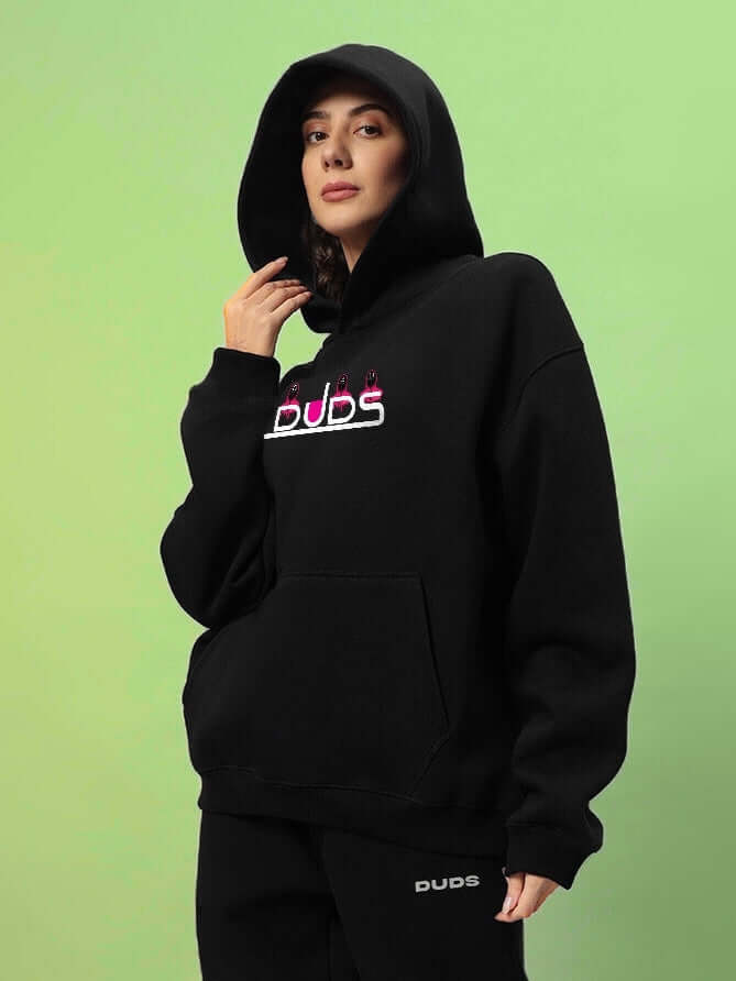 Woman wearing a black WOMENS SQUID GAME OVERSIZED HOODIE BLACK with minimalist design on green background.