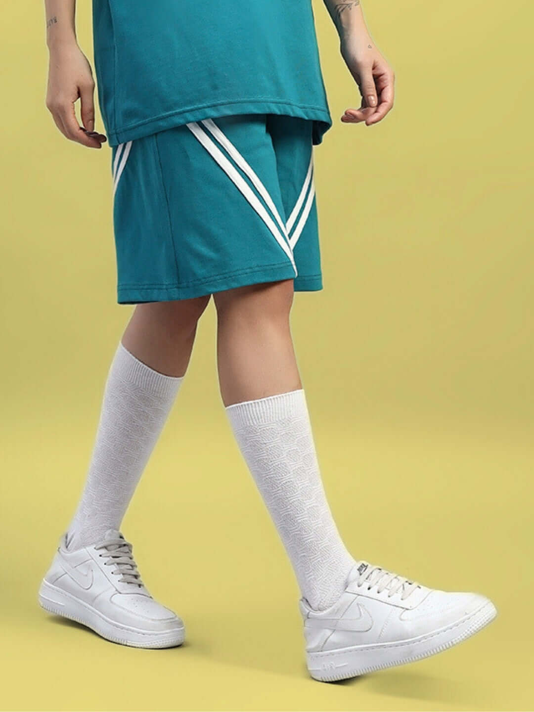 Model wearing WOMEN'S STAR JUMPER REGULAR FIT SHORTS TEAL with white shoes and socks on a yellow background.