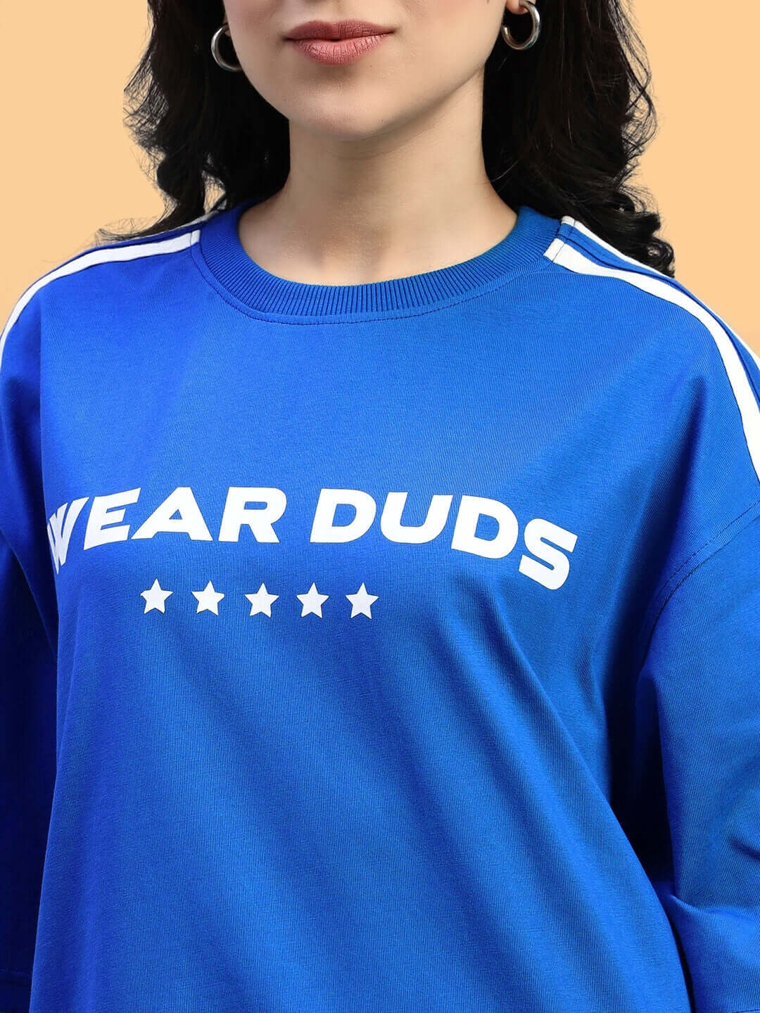 Woman wearing blue oversized t-shirt from WOMENS STAR JUMPER SUMMER CO-ORD SET BLUE, featuring stars and bold text, against a beige background.