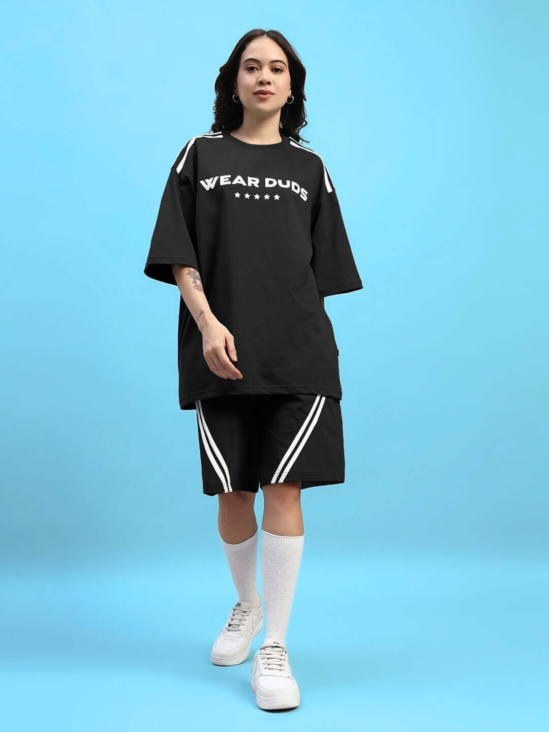 Woman wearing a black co-ord set with "Wear Dudes" text, consisting of an oversized top and shorts, against a blue background.