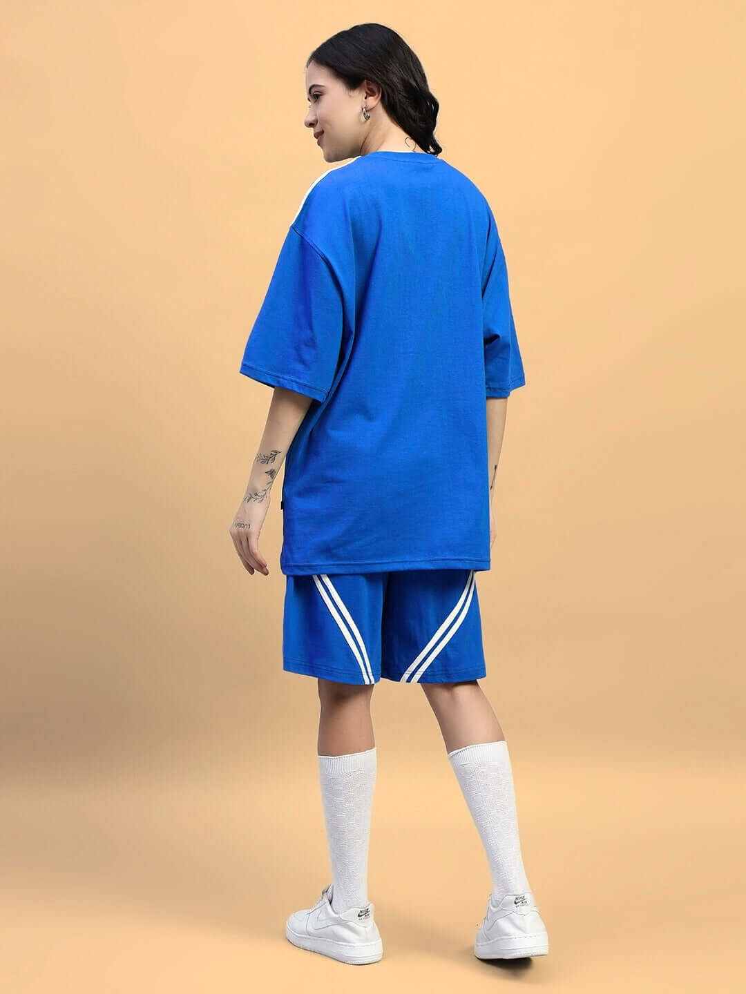 Woman wearing WOMENS STAR JUMPER SUMMER CO-ORD SET BLUE with oversized t-shirt and shorts, showing back view against a peach background.