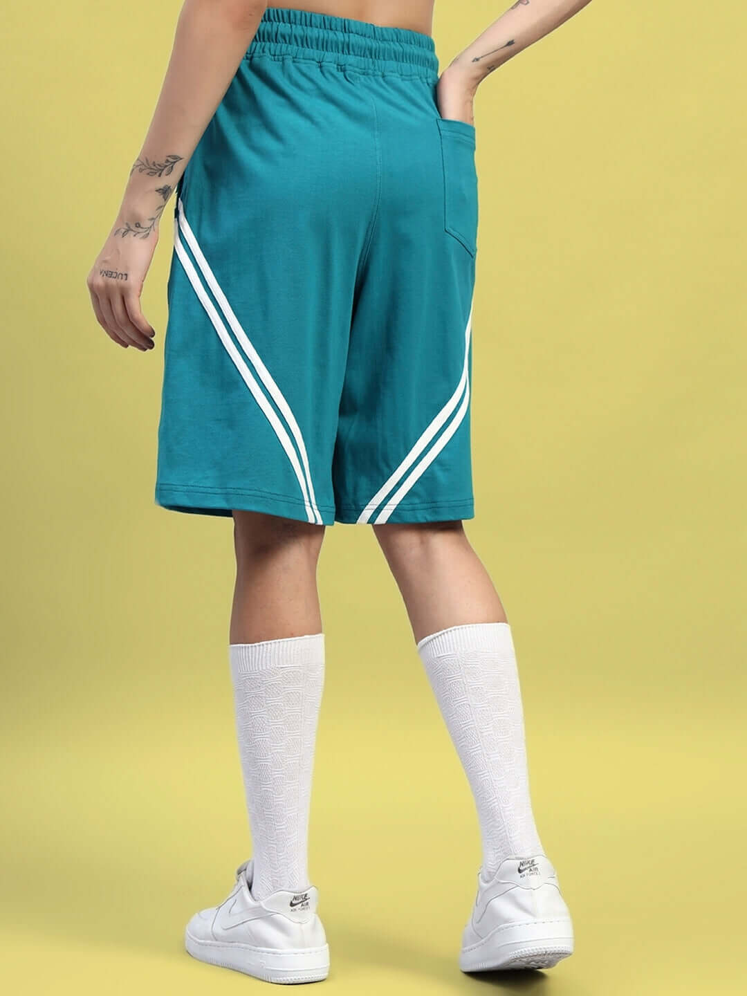 Back view of a model wearing teal shorts with white stripes, paired with white socks and shoes, on a yellow background.