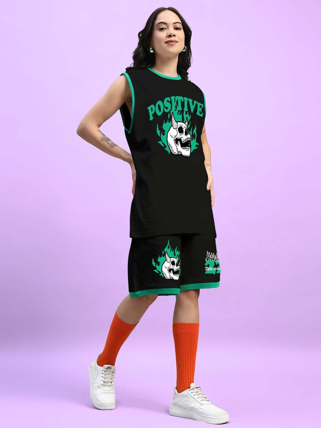 Woman wearing WOMENS STAY POSITIVE CO-ORD SET BLACK with skull graphic, matching shorts, orange socks, and white sneakers.