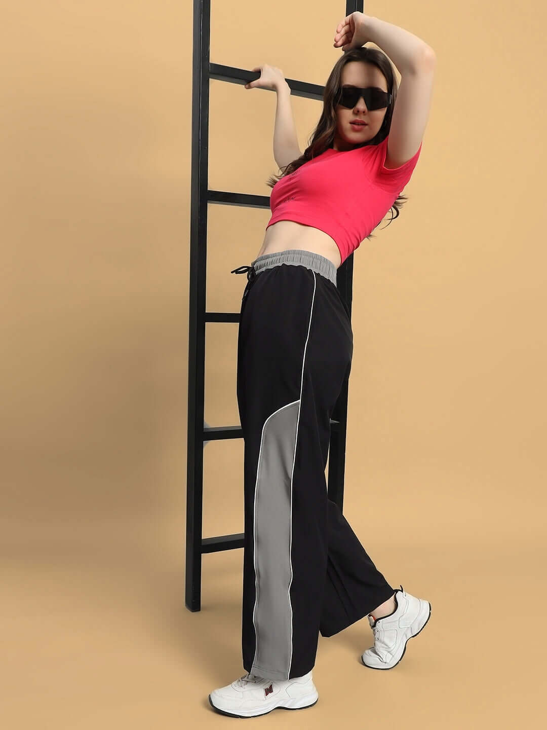 Woman wearing Women's Strom Relaxed Fit Cargo Pants in Black-Grey, styled with a pink top and sunglasses, striking a pose next to a ladder.