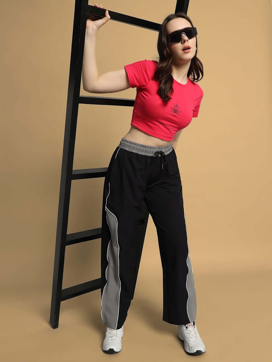 Model wearing Women's Strom Relaxed Fit Cargo Pants in black-grey with a pink crop top and sunglasses, leaning against a ladder.