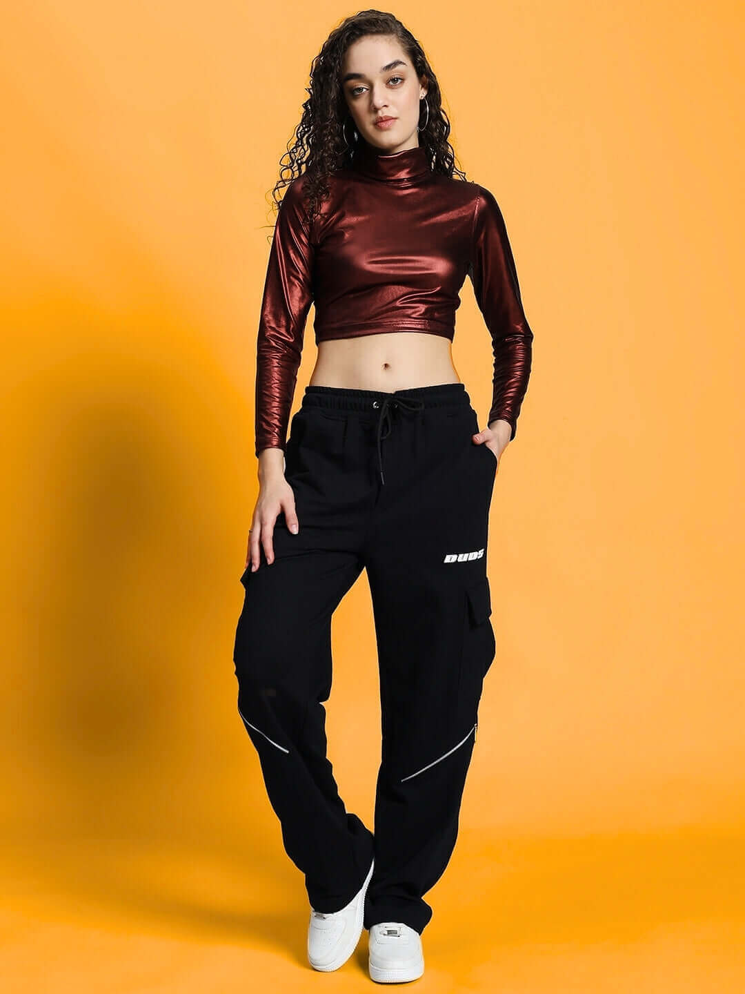 Woman wearing maroon metallic crop top with black pants, standing against a yellow background.