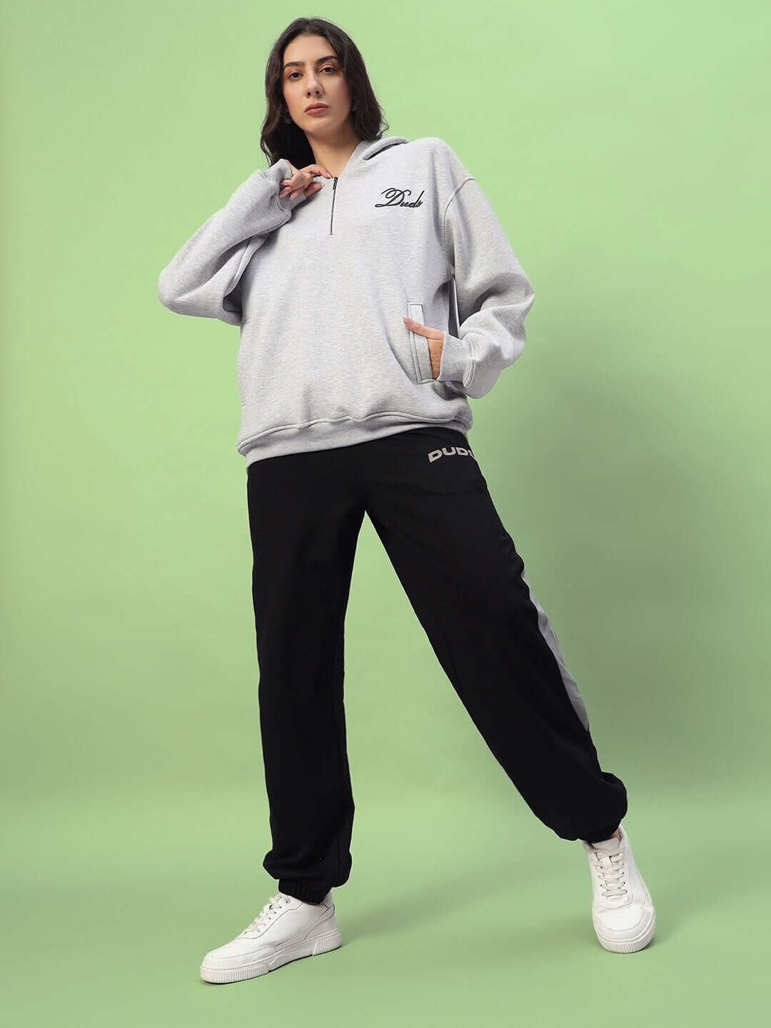 Woman wearing black-grey baggy joggers with a grey hoodie, standing on green background. Stylish and comfortable outfit for casual wear.