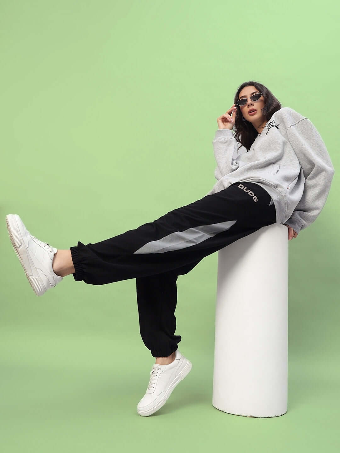 Model wearing DUDS Women's Trackpoint Baggy Joggers in black-grey, styled with a casual sweater and white sneakers on a green backdrop.