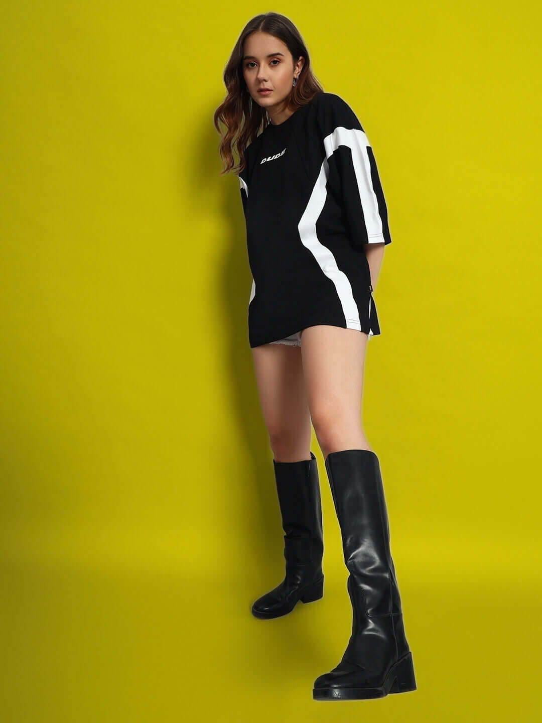 Model wearing women's oversized black-white t-shirt with premium cotton, styled with black boots against a yellow backdrop.