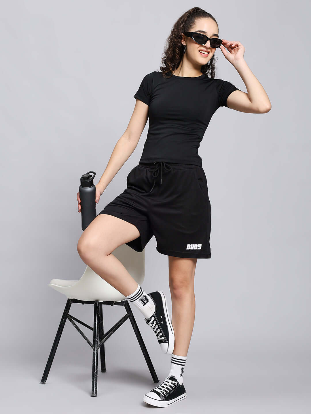 Woman in black tactical shorts holding a water bottle, wearing sunglasses and sneakers, posing casually on a white chair.